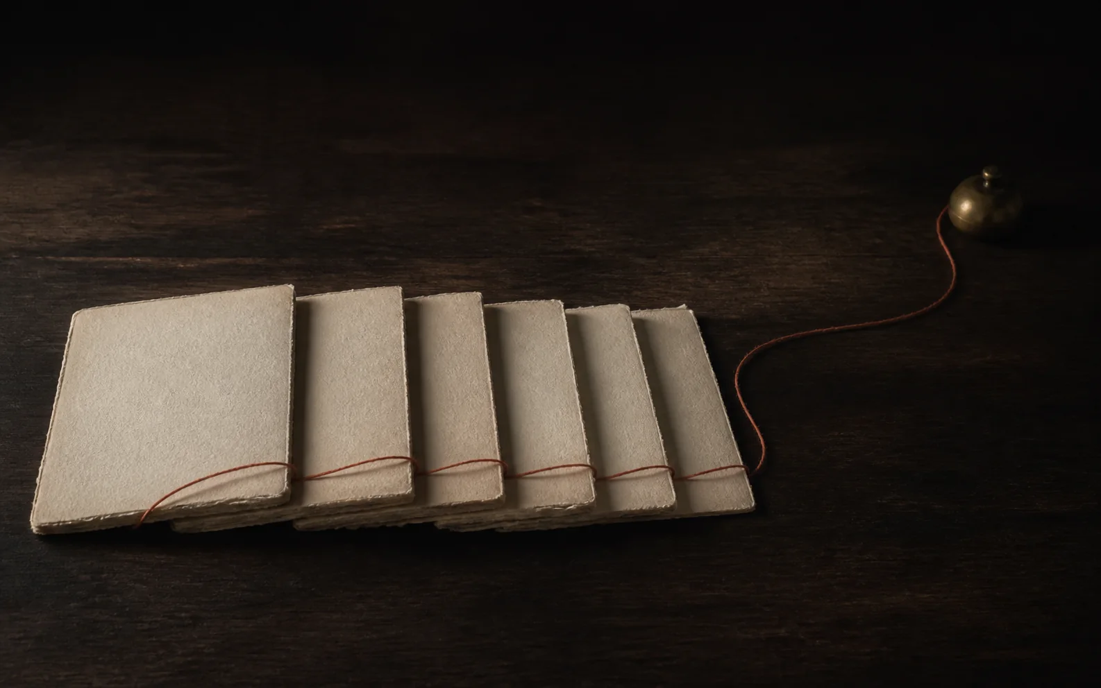 Six loose gatherings of blank handmade paper laid in a row on dark wood, a single terracotta thread running through all of them and away to a small brass bell.